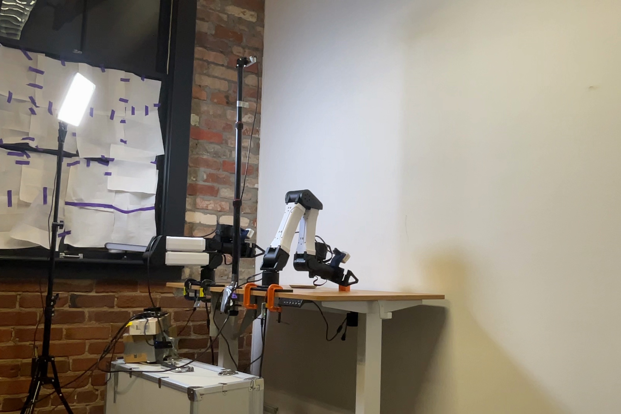 An I2RT YAM arm clamped to a wooden desk against a white wall, a camera on a pole behind it, a small orange cube on the table and a light panel on the left